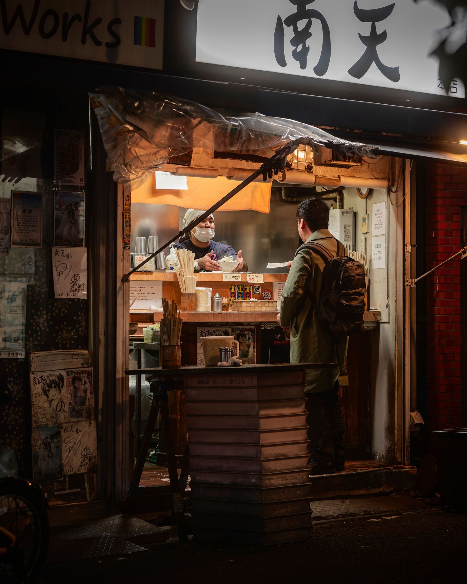 A customer orders at a warmly lit Japanese street food stall at night, with kanji signage glowing above.