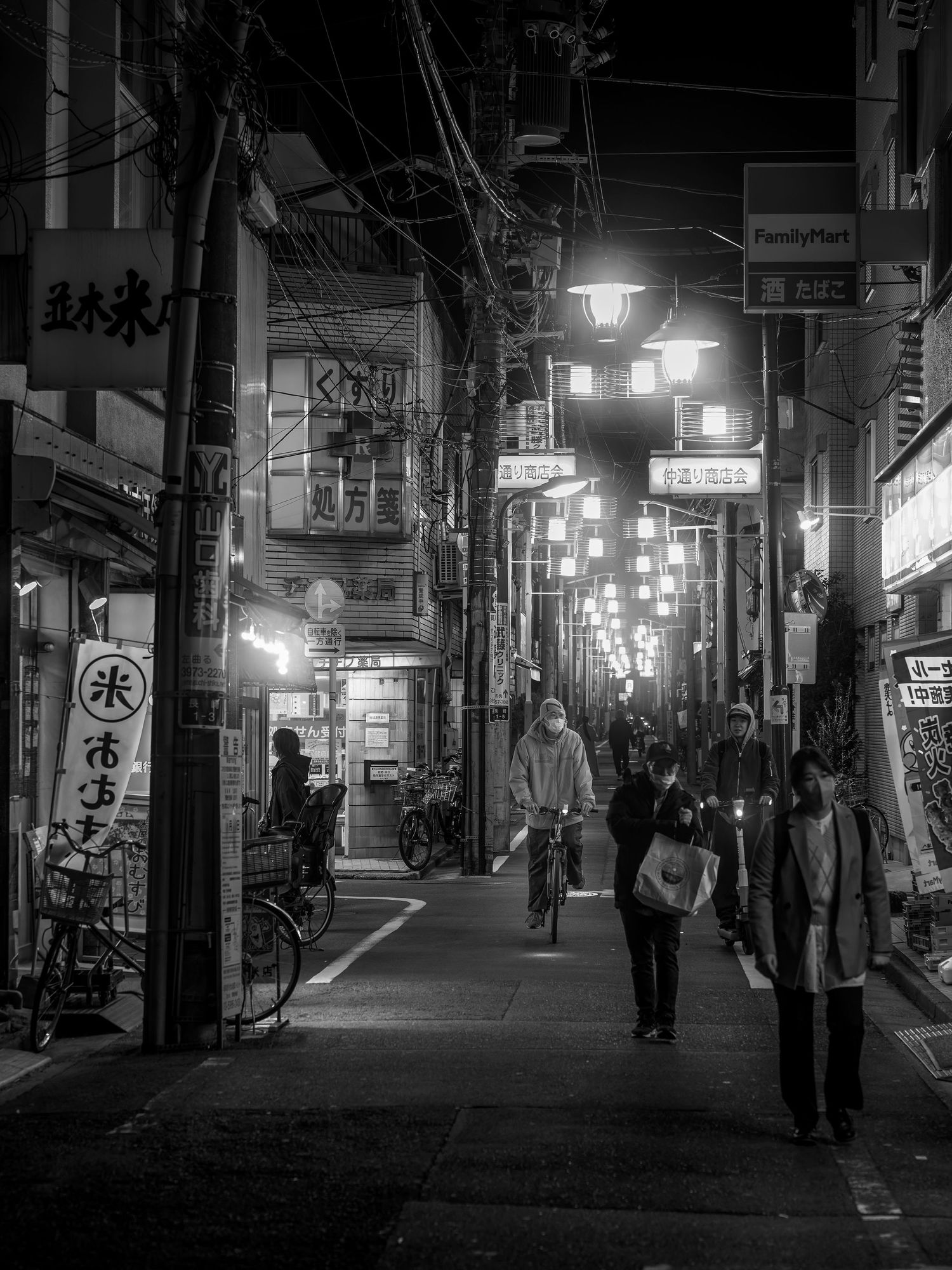 Black and white night street scene in Japan with pedestrians, cyclist, glowing lanterns, and FamilyMart sign.
