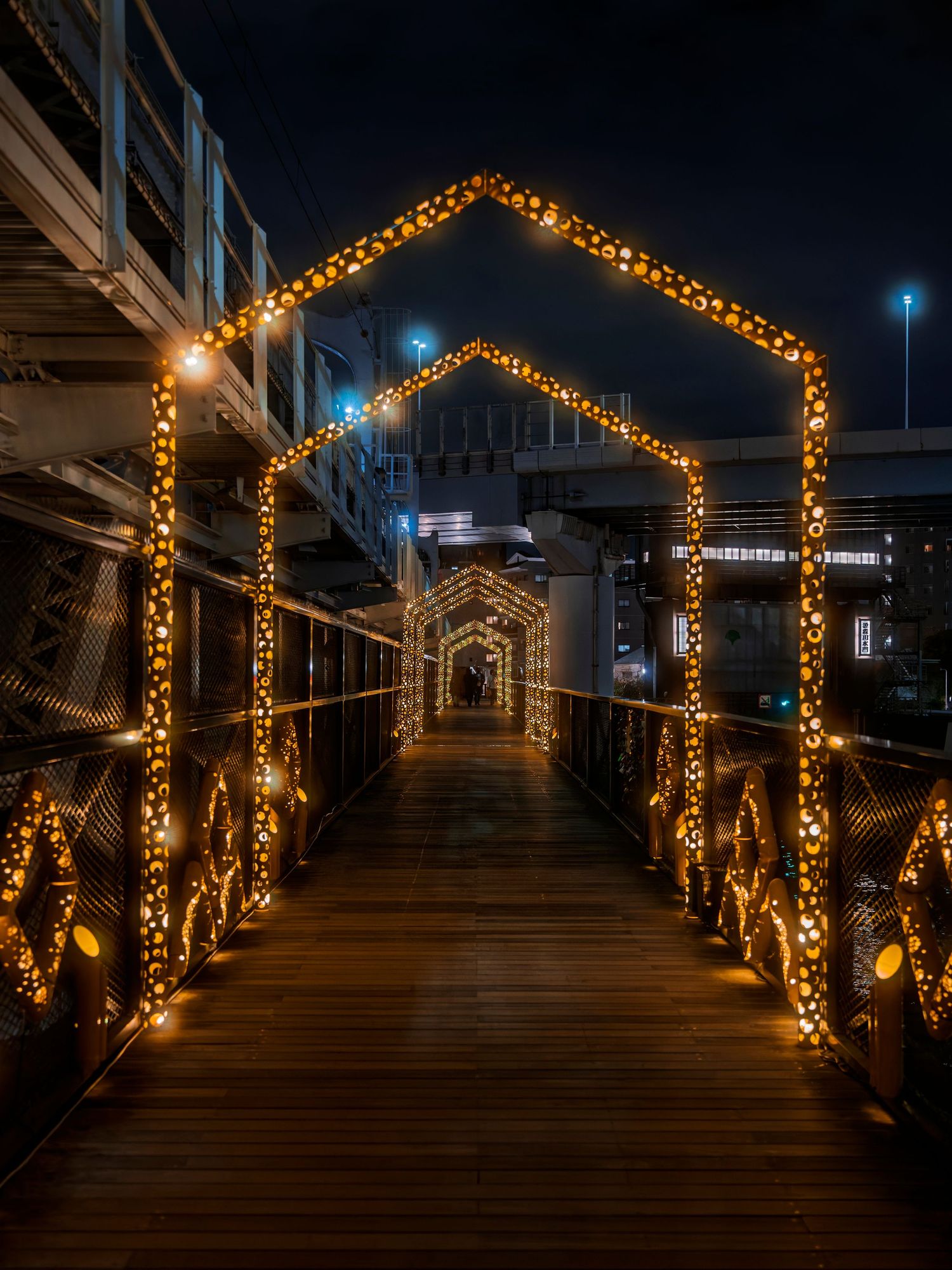 A wooden bridge walkway illuminated by warm golden fairy lights forming archways at night.
