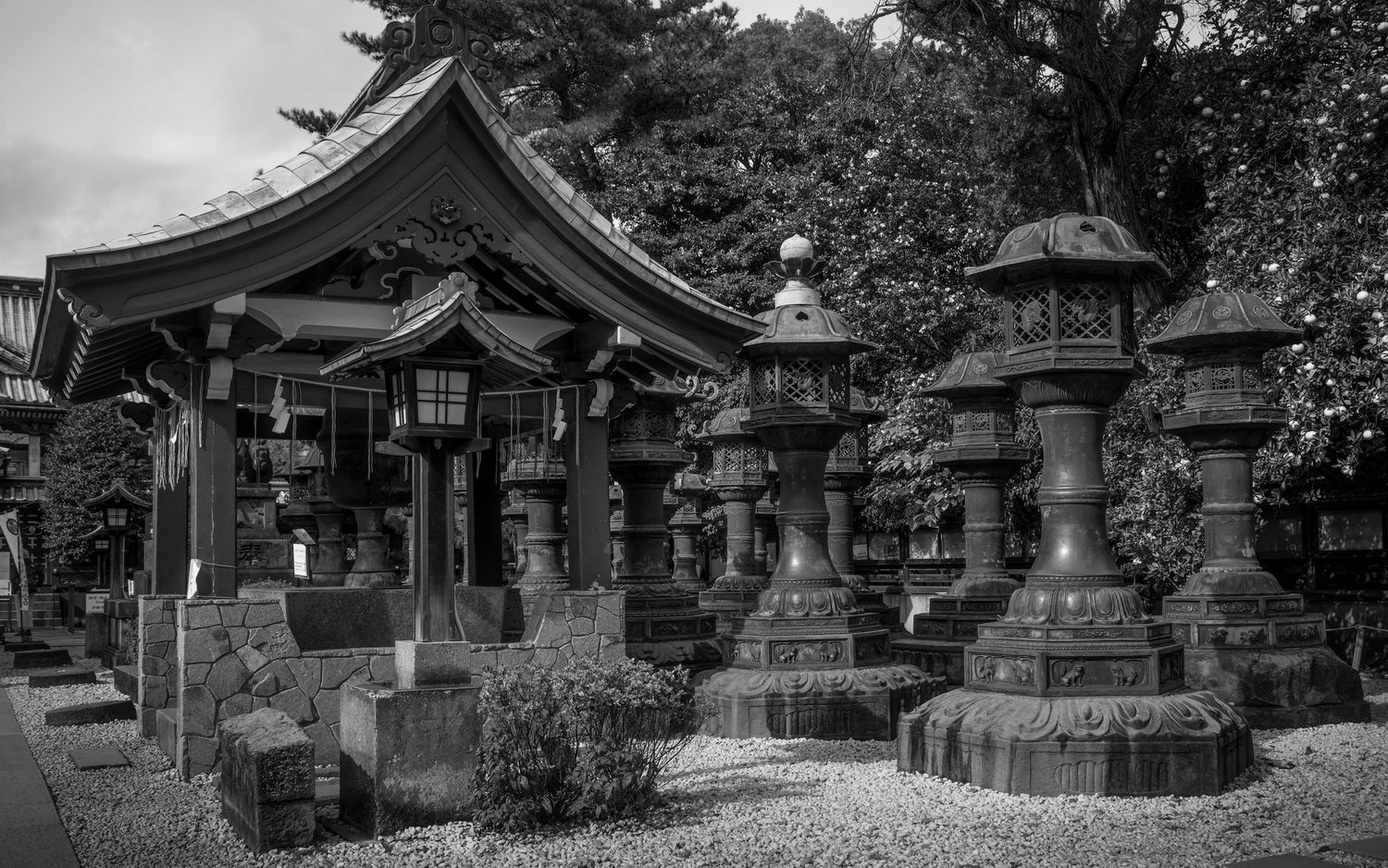 Black and white photo of a traditional Japanese shrine with stone lanterns and pagoda architecture surrounded by trees.