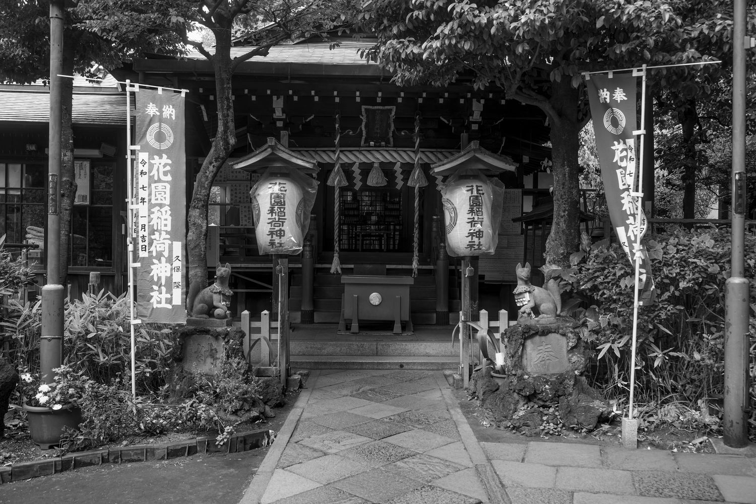 Black and white photo of a traditional Japanese shrine entrance with lanterns, banners, and stone guardians lining a stone path.
