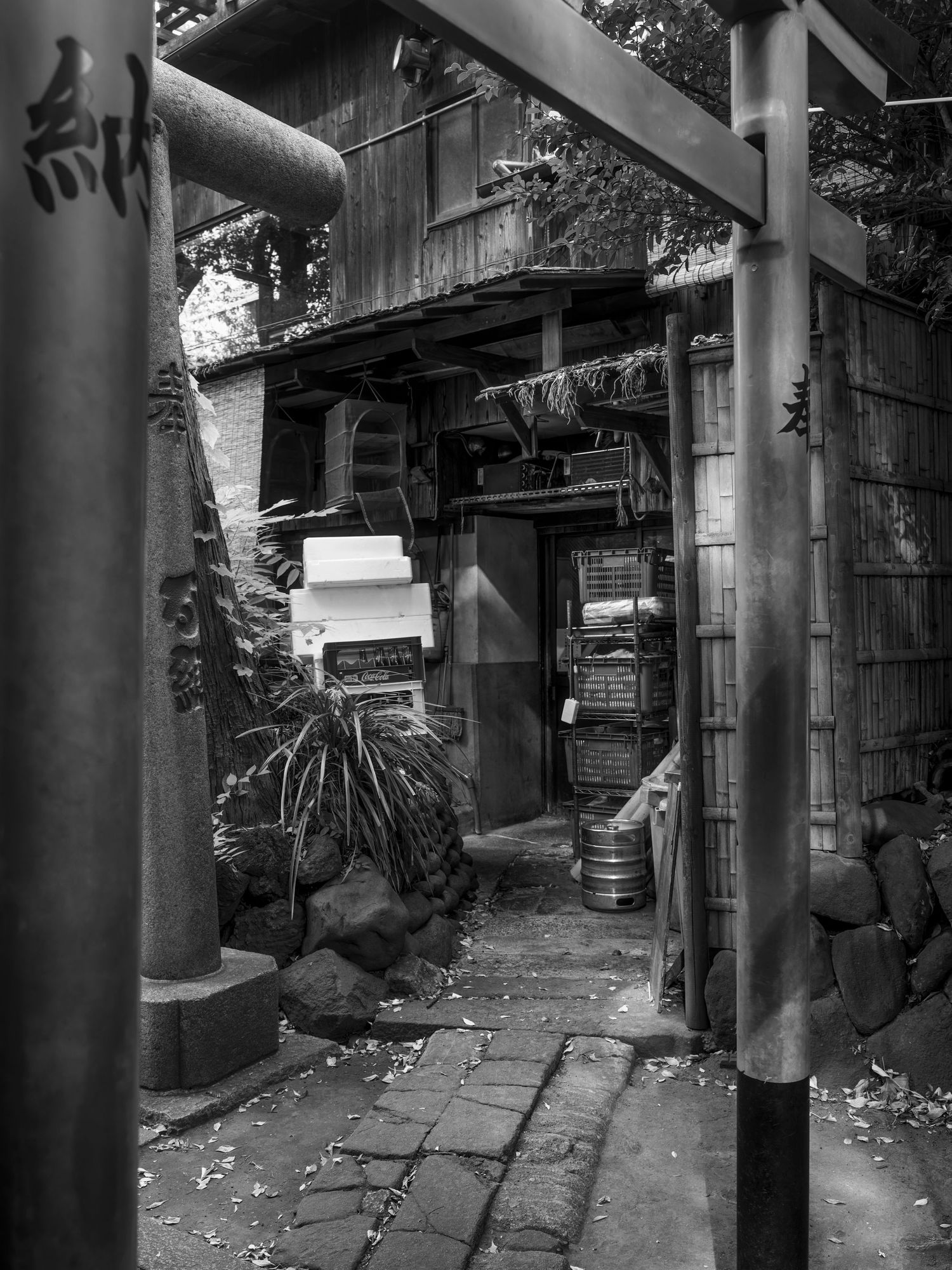Black and white photo of a rustic Japanese alleyway with torii gate, stone path, and weathered wooden structures.