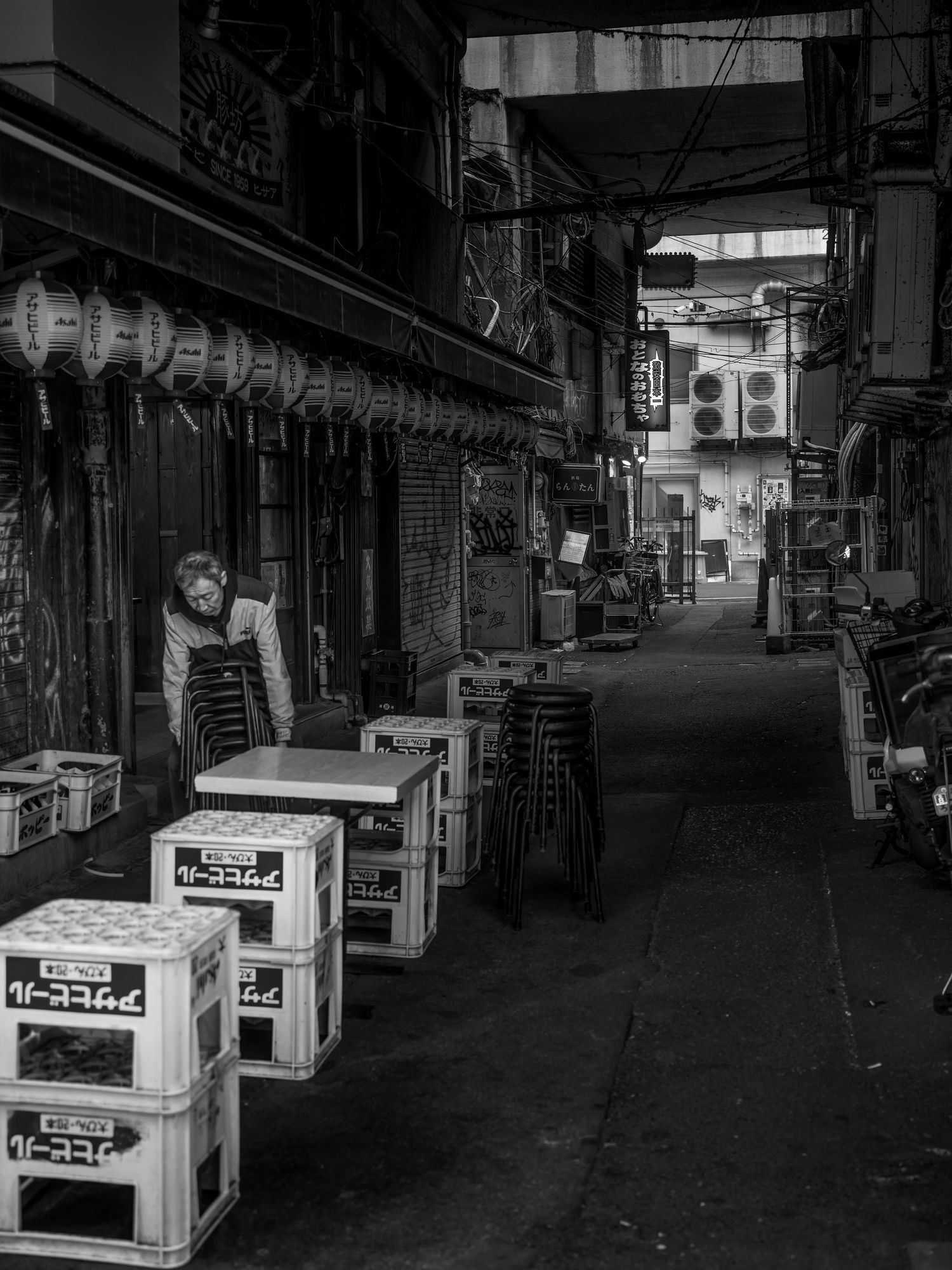 Black and white photo of a narrow Japanese alley with stacked chairs, crates, and a worker outside a closed restaurant.