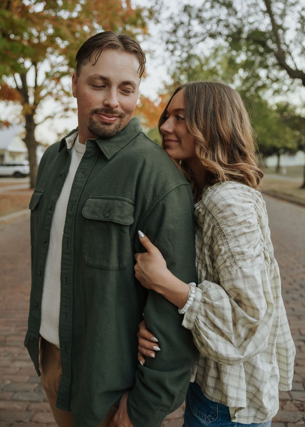A woman in a plaid shirt embraces a man in a green jacket while standing on a brick path in fall.