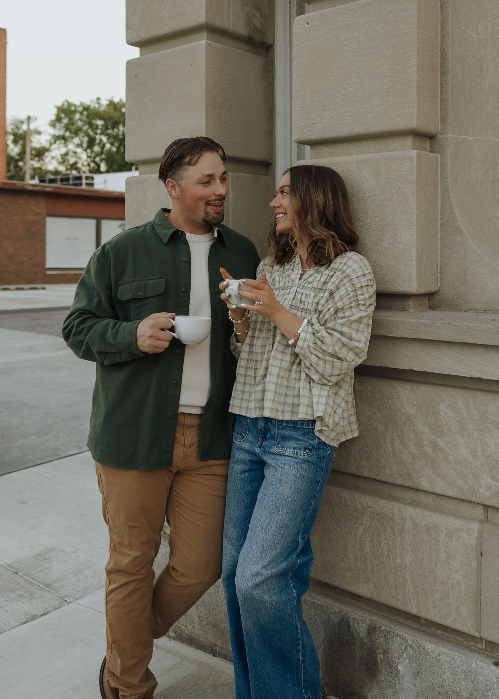 Smiling couple with coffee mugs leaning against stone building column on city street.