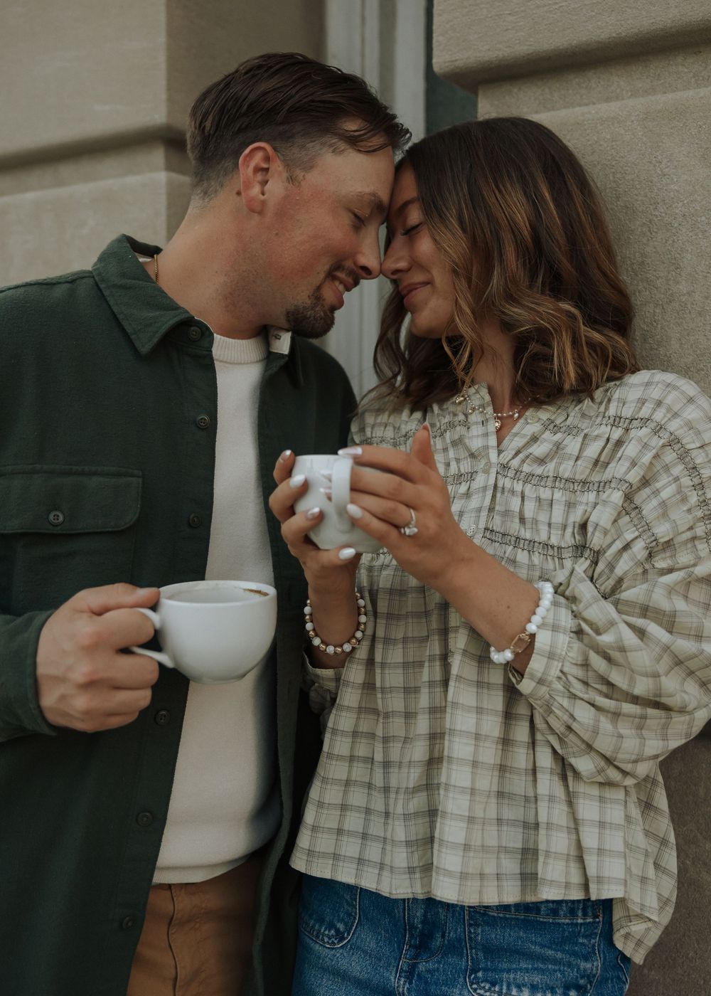 Couple touching foreheads romantically while holding white coffee mugs against stone wall.