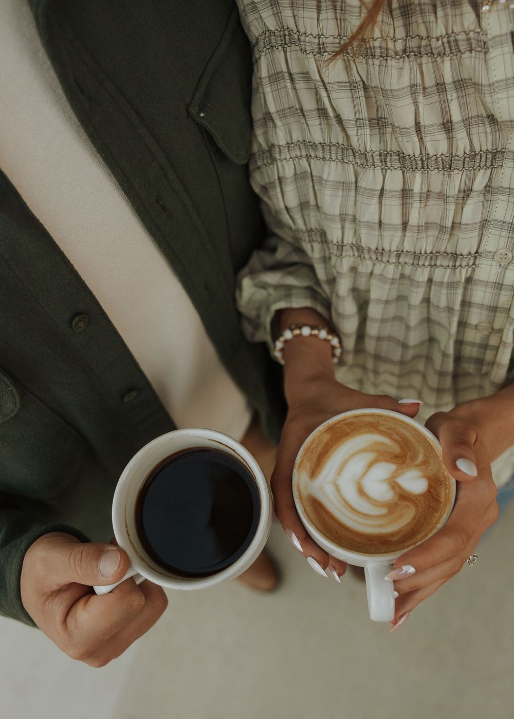 Two people holding coffee cups, one black coffee and one latte with leaf art, wearing casual autumn outfits.
