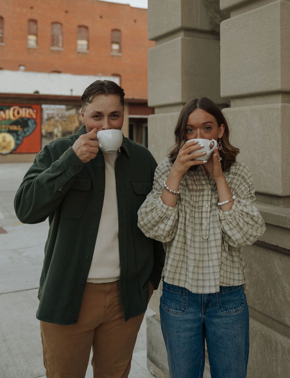 A couple sipping from white coffee mugs outdoors on a city sidewalk in coordinating green and plaid outfits.