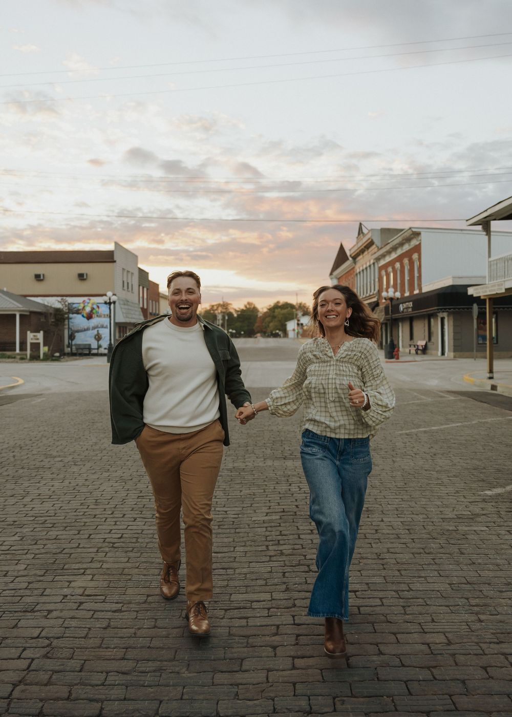 A happy couple holding hands and running through a brick town square at sunset with colorful sky.