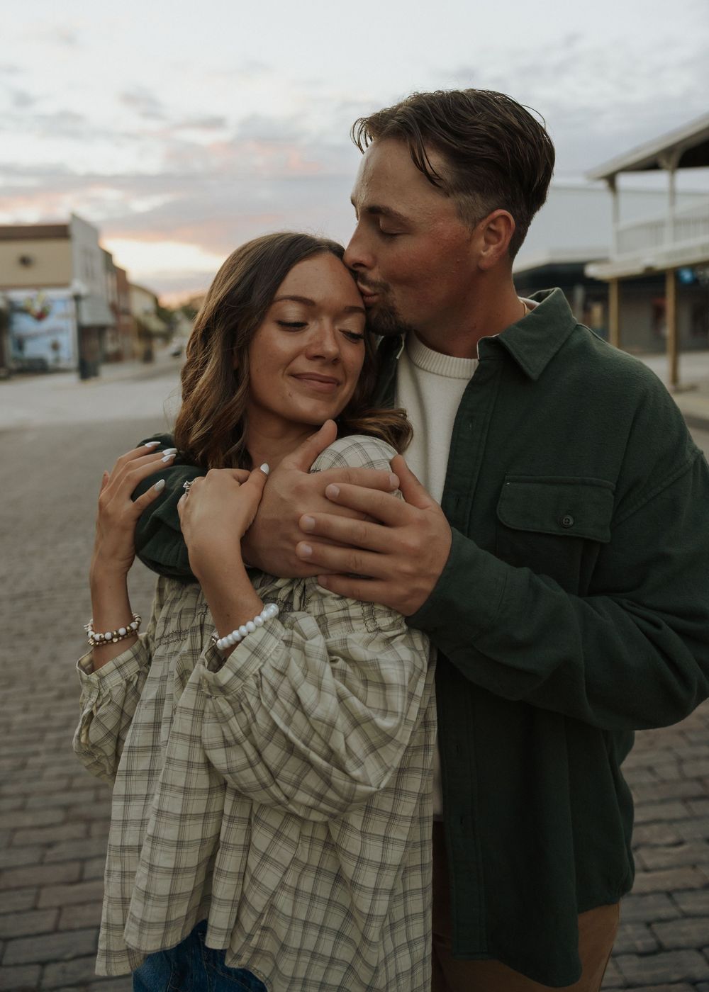 A man in a green jacket tenderly kisses a smiling woman on the forehead during a romantic outdoor shoot.