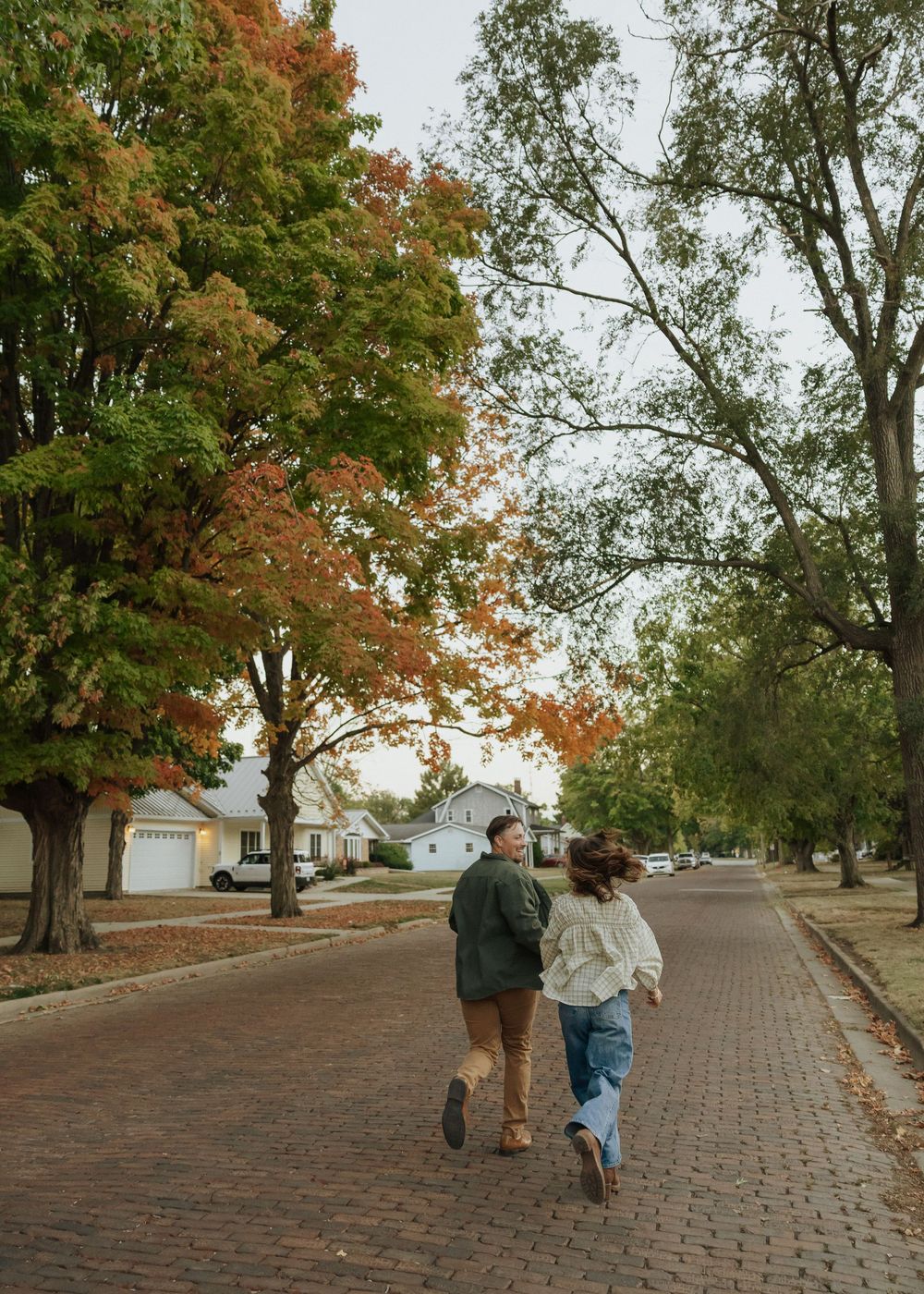 A couple runs hand-in-hand down a brick tree-lined street surrounded by colorful autumn foliage.
