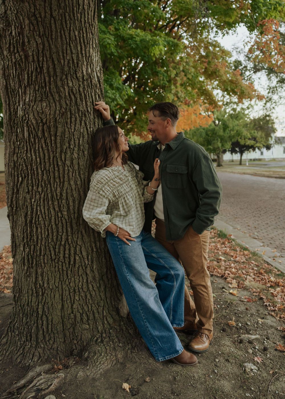 Couple leaning against a large tree trunk on a fall day, smiling at each other affectionately.