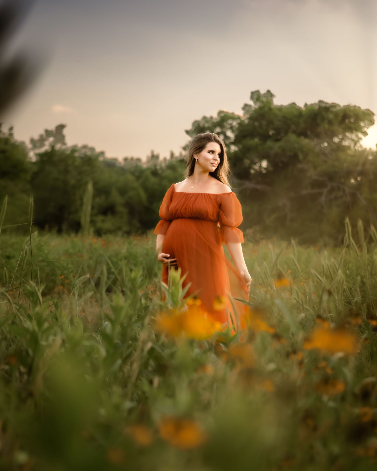 A pregnant woman in an orange dress stands thoughtfully in a wildflower meadow at golden hour in Fort Worth, TX
