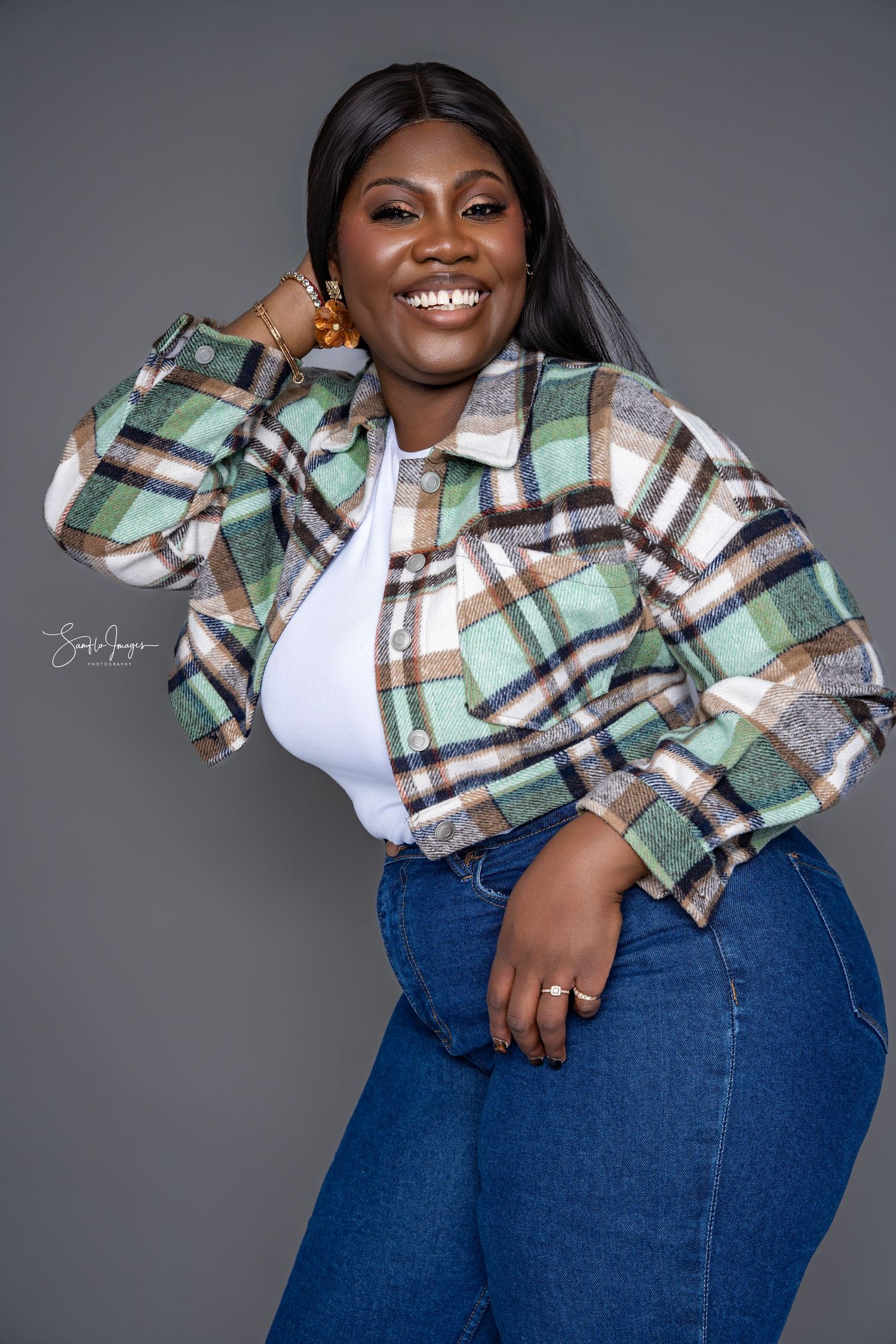 A person wearing a plaid cropped jacket and blue jeans poses confidently against a grey studio backdrop.