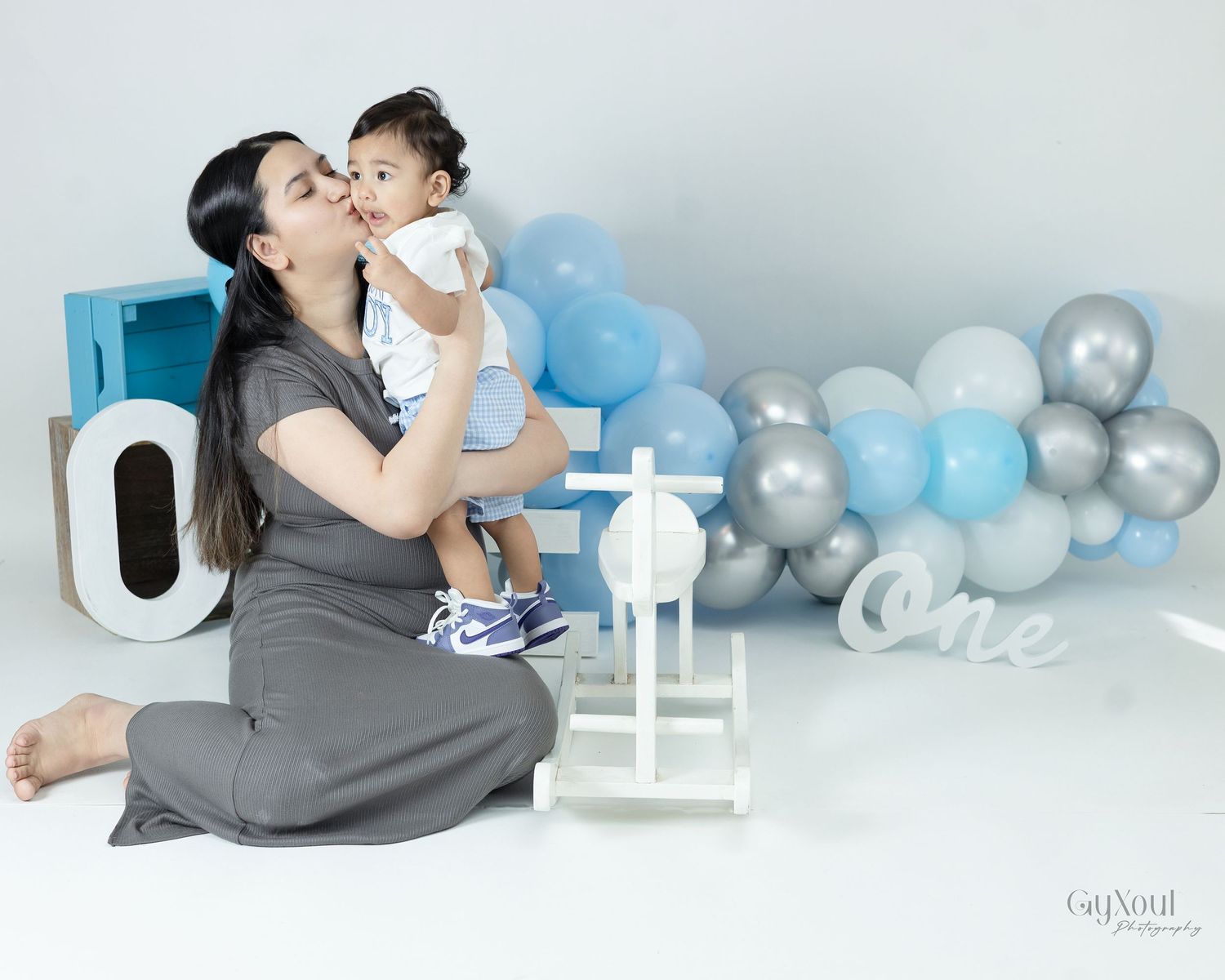 A baby and adult sit together in a studio setting with blue and silver balloons and decorative elements in the background.