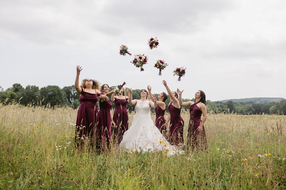 Bridesmaids in burgundy dresses throw bouquets in air for wedding photos on a cloudy day in a country field. Seacoast wedding photographer