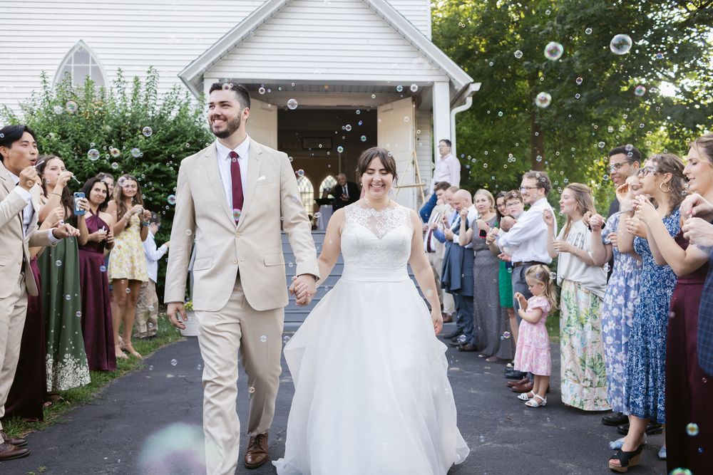 A bride and groom exit a white church as their wedding guests blow bubbles for their wedding exit in Bradford, Essex County MA