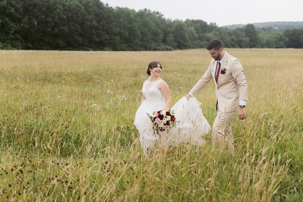 Groom in a tan suit holds his bride's dress walking in a field on a cloudy day on their wedding. Western, Lenox MA wedding photographer