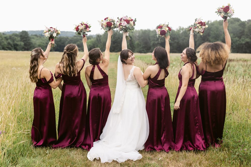 Bridal party wearing cranberry dresses raises their bouquets in celebration for wedding photos. Meredith, Concord wedding photographer