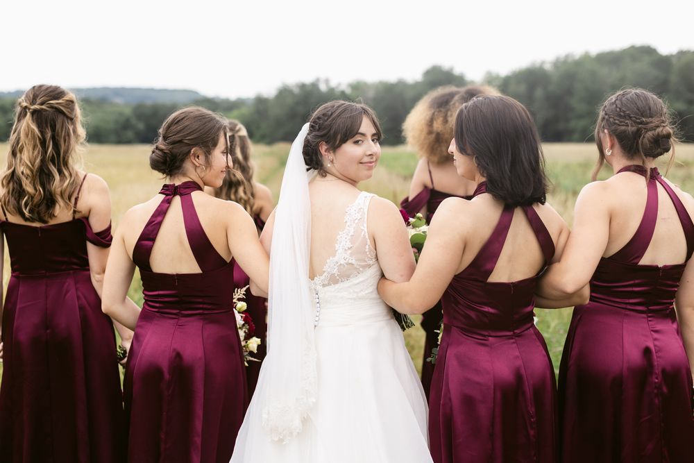 A group of bridesmaids in burgundy dresses stand together in a green field for wedding photos. Monadnock Region wedding photographer