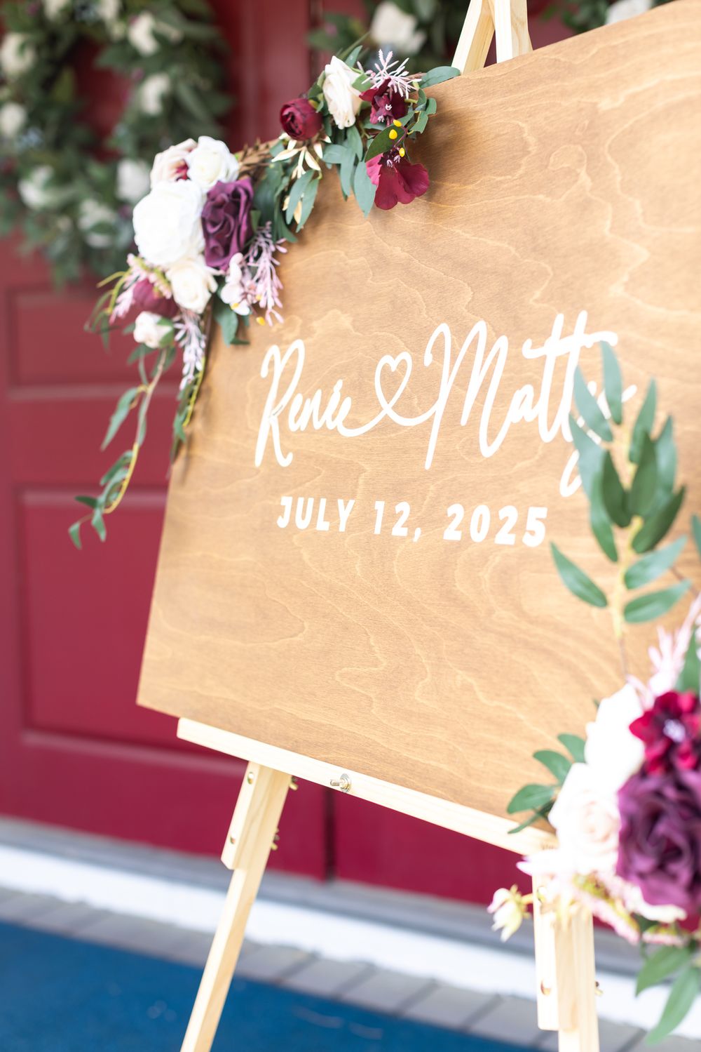 Wooden wedding welcome sign decorated with burgandy and white flowers displays names outside church venue in Bradford, MA for a wedding