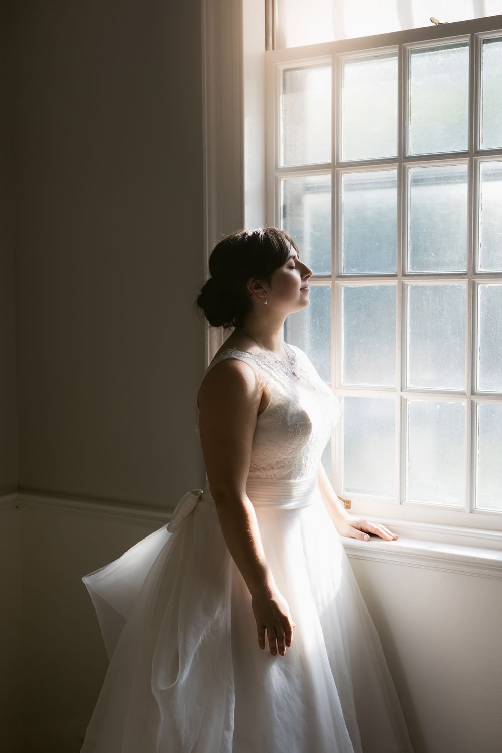A bride in a wedding dress stands by a window in soft natural light creating a romantic silhouette on her wedding day in Bradford, MA.