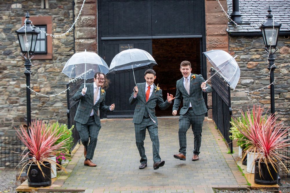 Three groomsmen in grey suits jump playfully with clear bubble umbrellas outside rustic stone wedding venue.