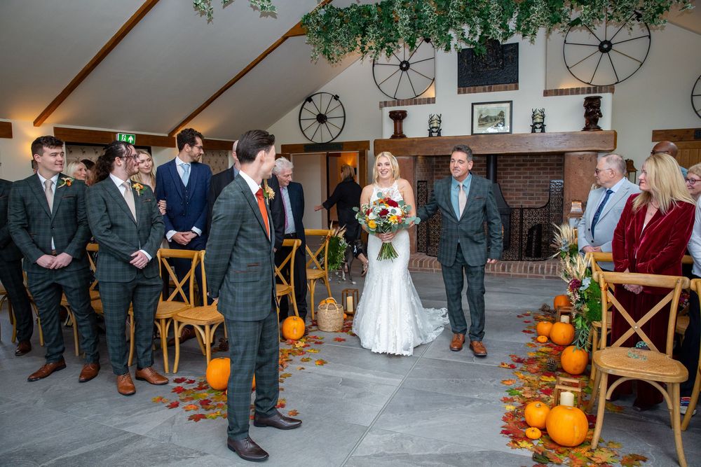 Bride in fitted lace gown walks down autumn-decorated aisle with pumpkins past groomsmen in tweed suits.