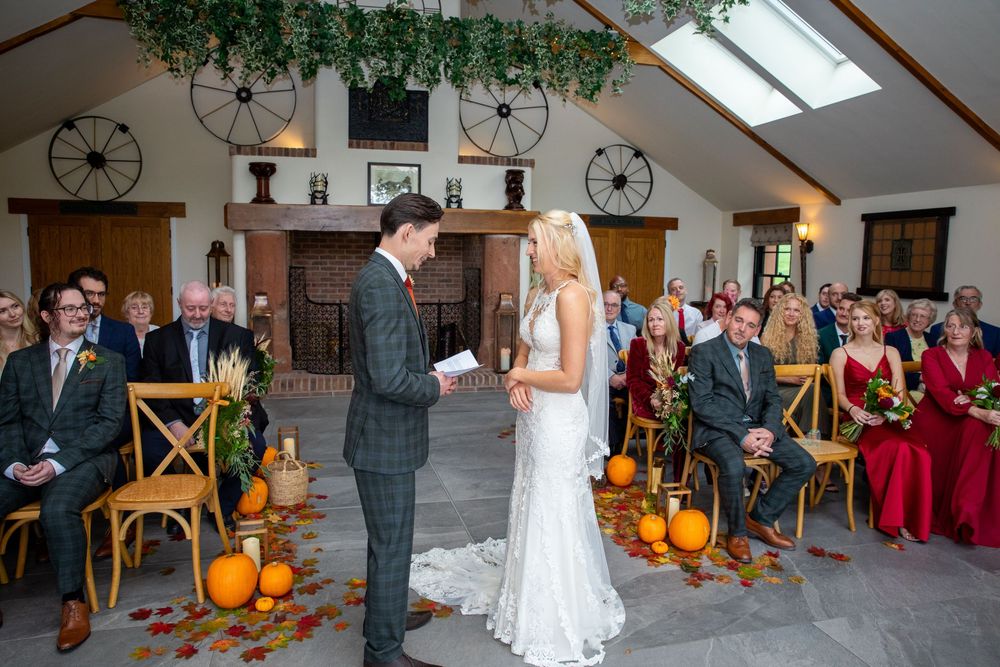 Groom reads vows to bride in lace dress during rustic autumn wedding ceremony decorated with pumpkins and leaves.