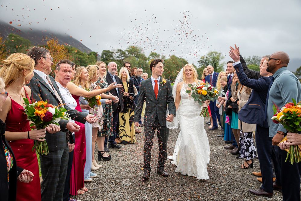 Wedding couple walks through confetti shower surrounded by guests at scenic outdoor celebration.