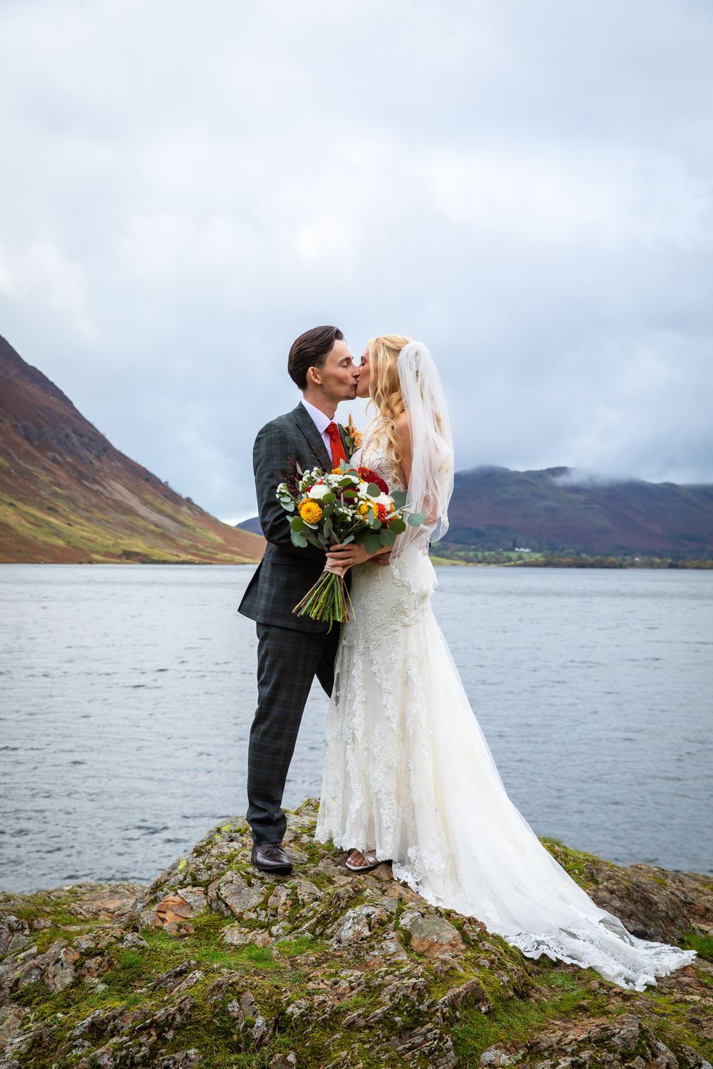Bride and groom share romantic lakeside kiss on rocky outcrop with misty mountains behind.