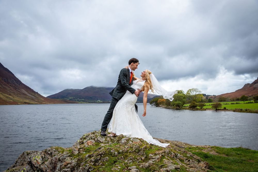 Groom dips bride on lakeside rock with dramatic cloudy mountain landscape in background.