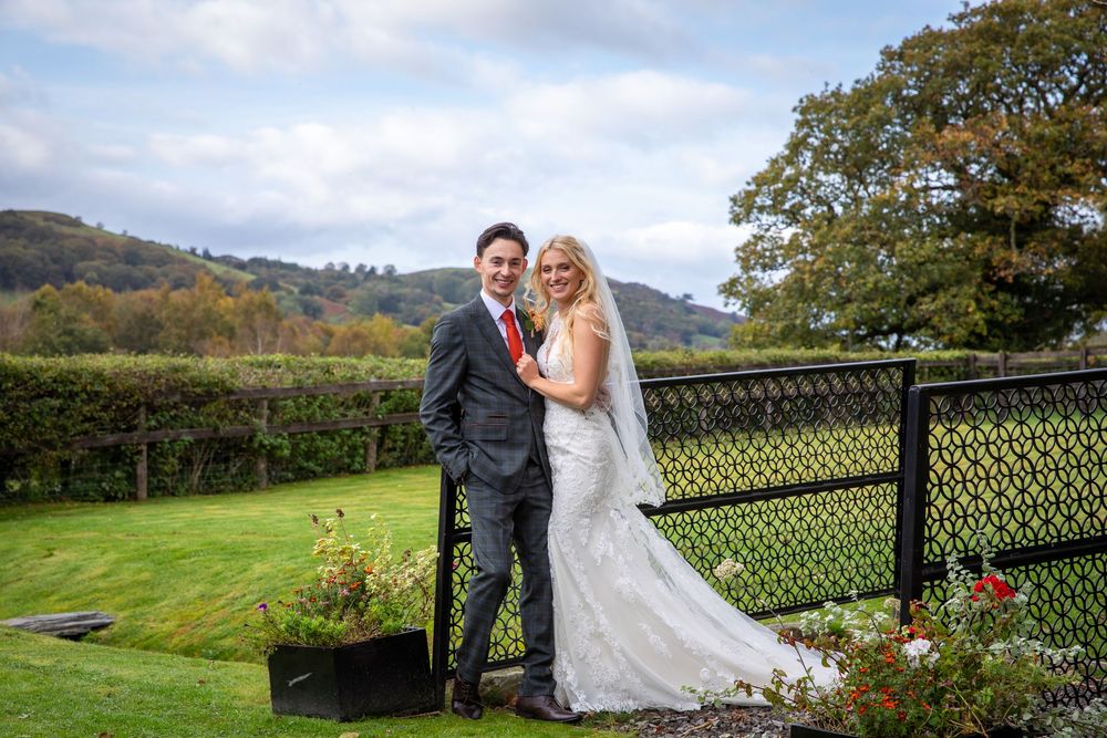 Smiling newlyweds pose by ornate garden fence with autumn hills and blue sky backdrop.