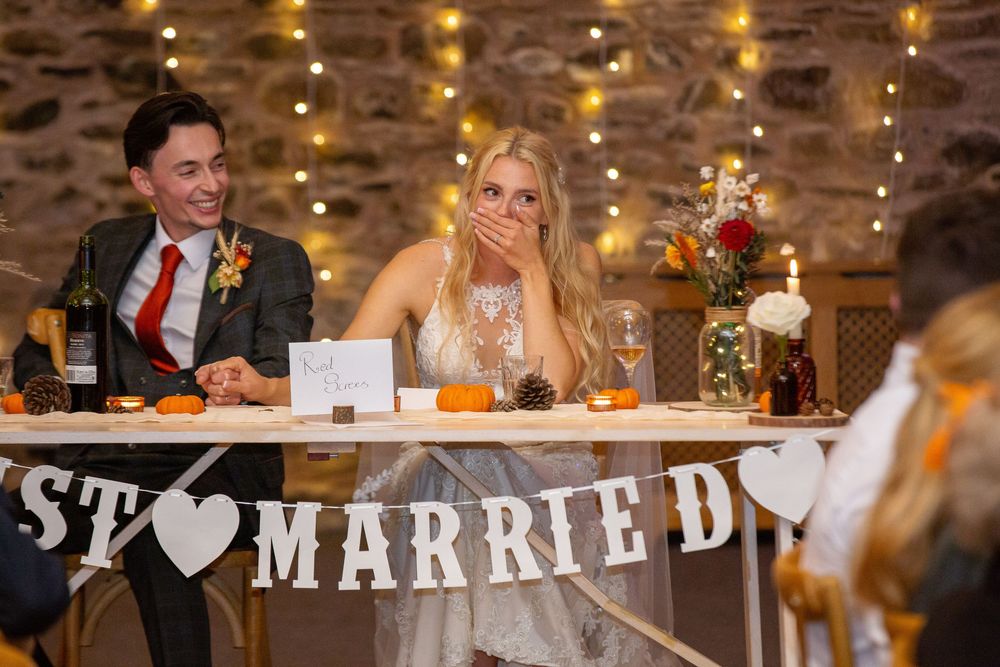 Bride laughs at reception table beneath rustic Just Married banner with glowing fairy lights.