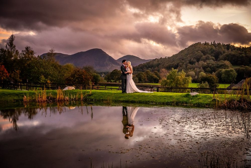 Bride and groom embrace at golden sunset reflected in still pond with mountain scenery.