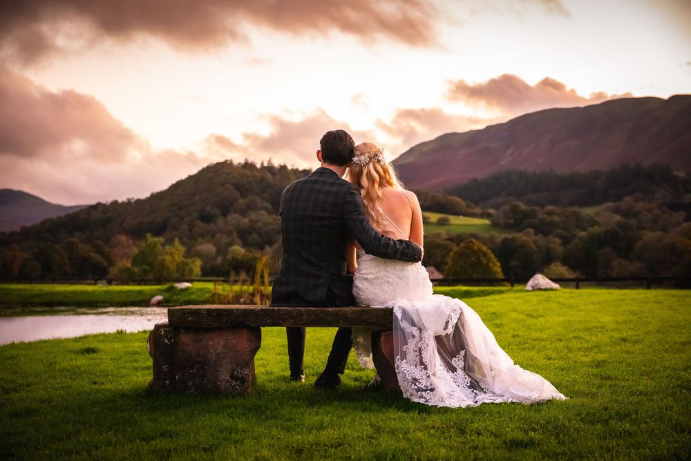 Newlyweds sit on rustic bench admiring dramatic mountain sunset landscape by tranquil lake.