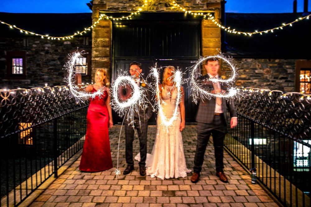 Wedding party writes LOVE with sparklers at night beneath fairy-lit stone archway entrance.