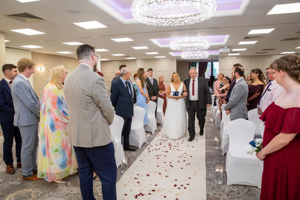 Bride walking down the aisle with escort in elegant chandelier-lit wedding ceremony venue.