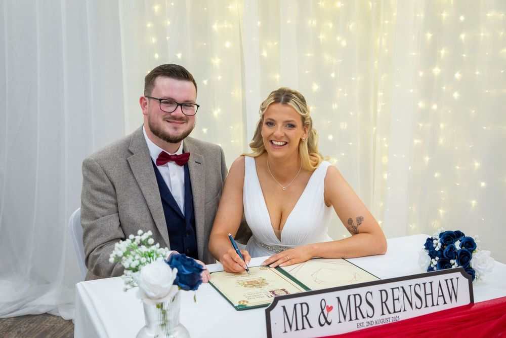 Mr and Mrs Renshaw smile while signing wedding register at decorated table with fairy light backdrop.