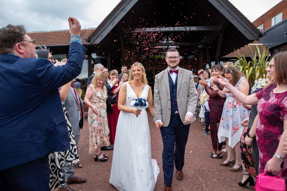 Newlyweds walk through confetti shower outside hotel venue surrounded by cheering wedding guests.