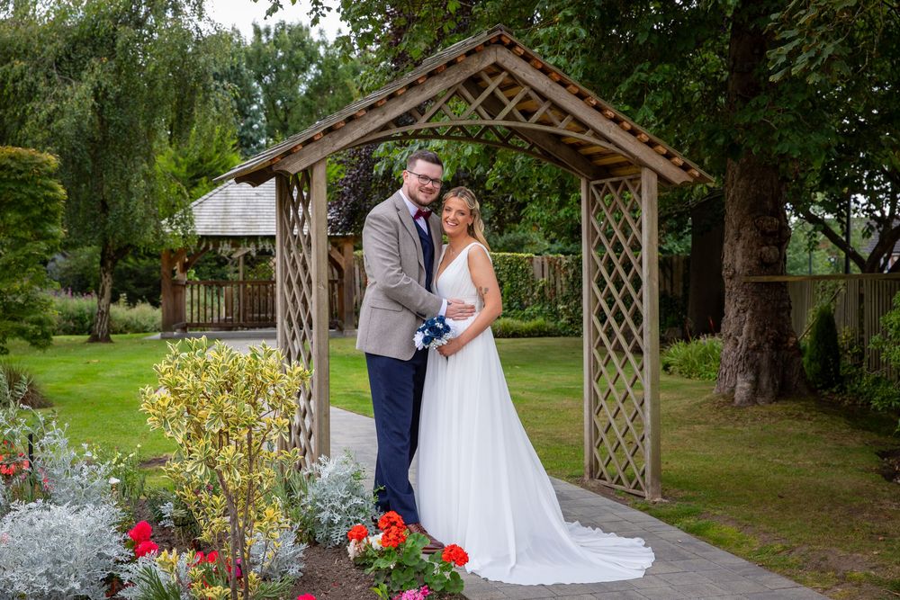 Bride and groom pose romantically beneath wooden garden arch surrounded by colorful flowers and greenery.