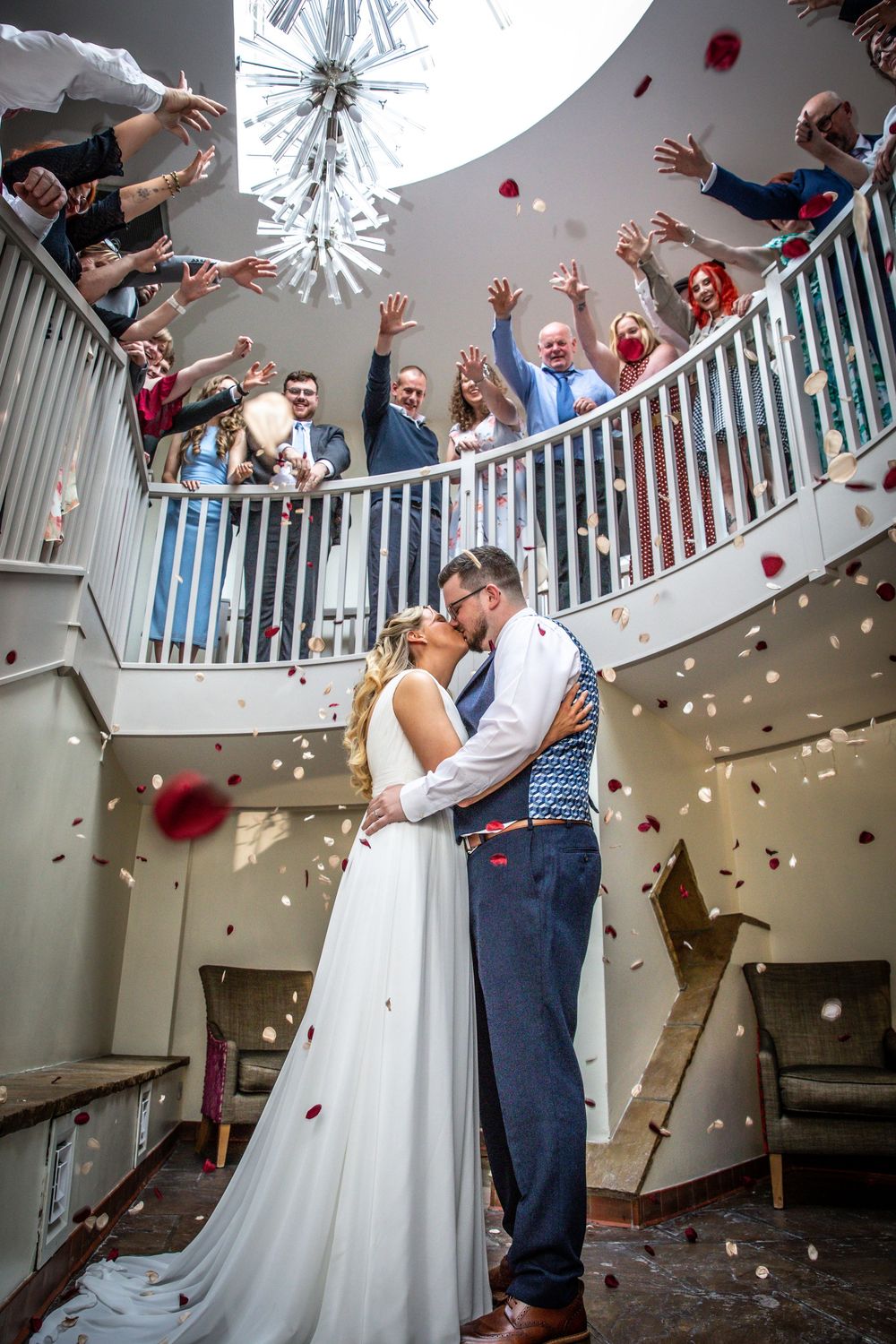 Bride and groom share first kiss under staircase as guests throw rose petals from balcony above.