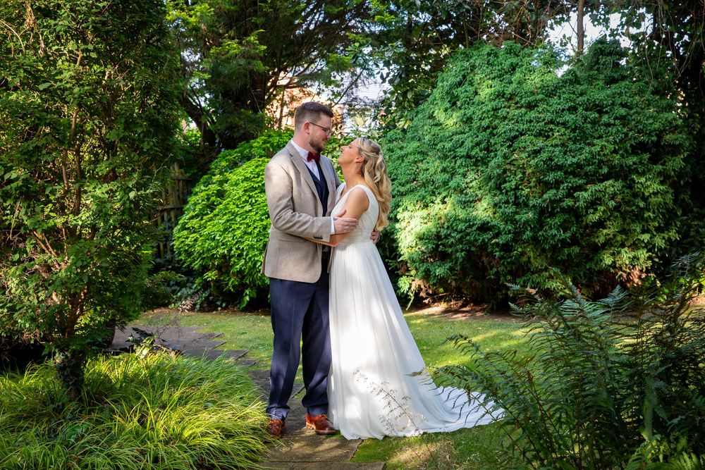 Bride and groom share tender moment surrounded by lush green garden foliage on wedding day.