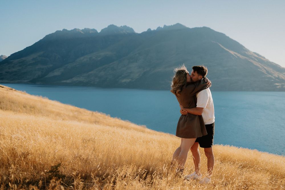 A couple embraces on a golden hillside overlooking a pristine mountain lake on a sunny afternoon.