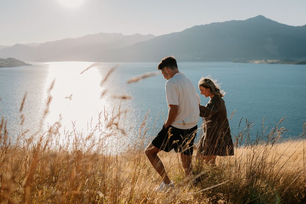 Two people sit together on a grassy hillside overlooking a scenic mountain lake on a bright sunny day.