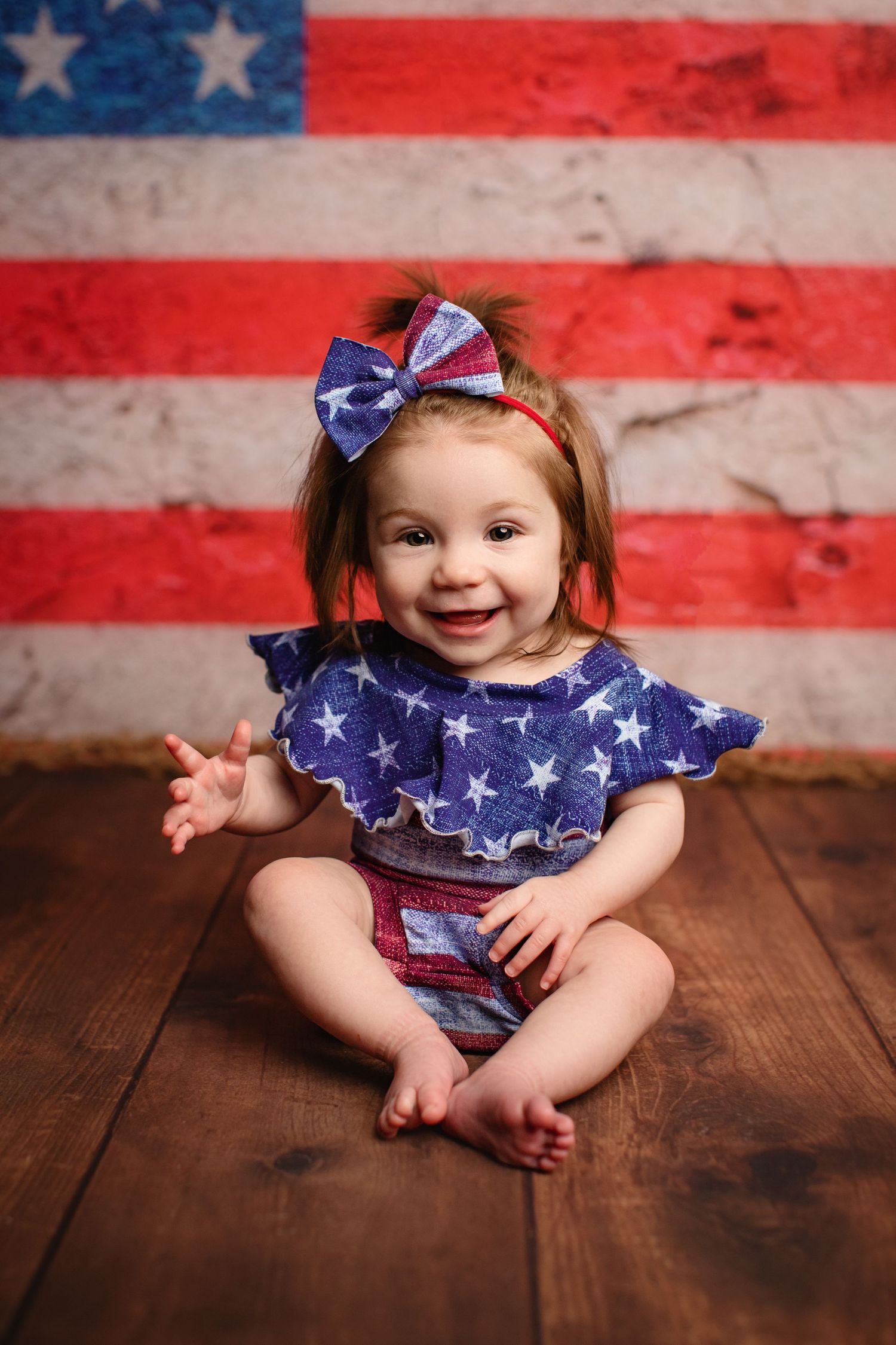 Patriotic baby photography series with American flag backdrop and blue star outfit on wooden floor.