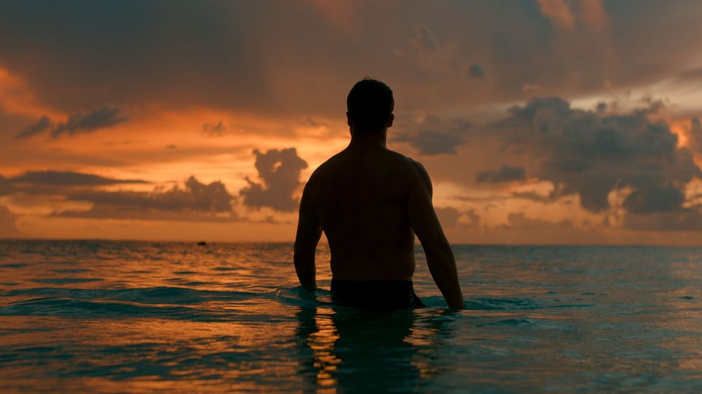 A silhouetted figure stands in calm ocean waters against a dramatic orange and purple sunset sky.