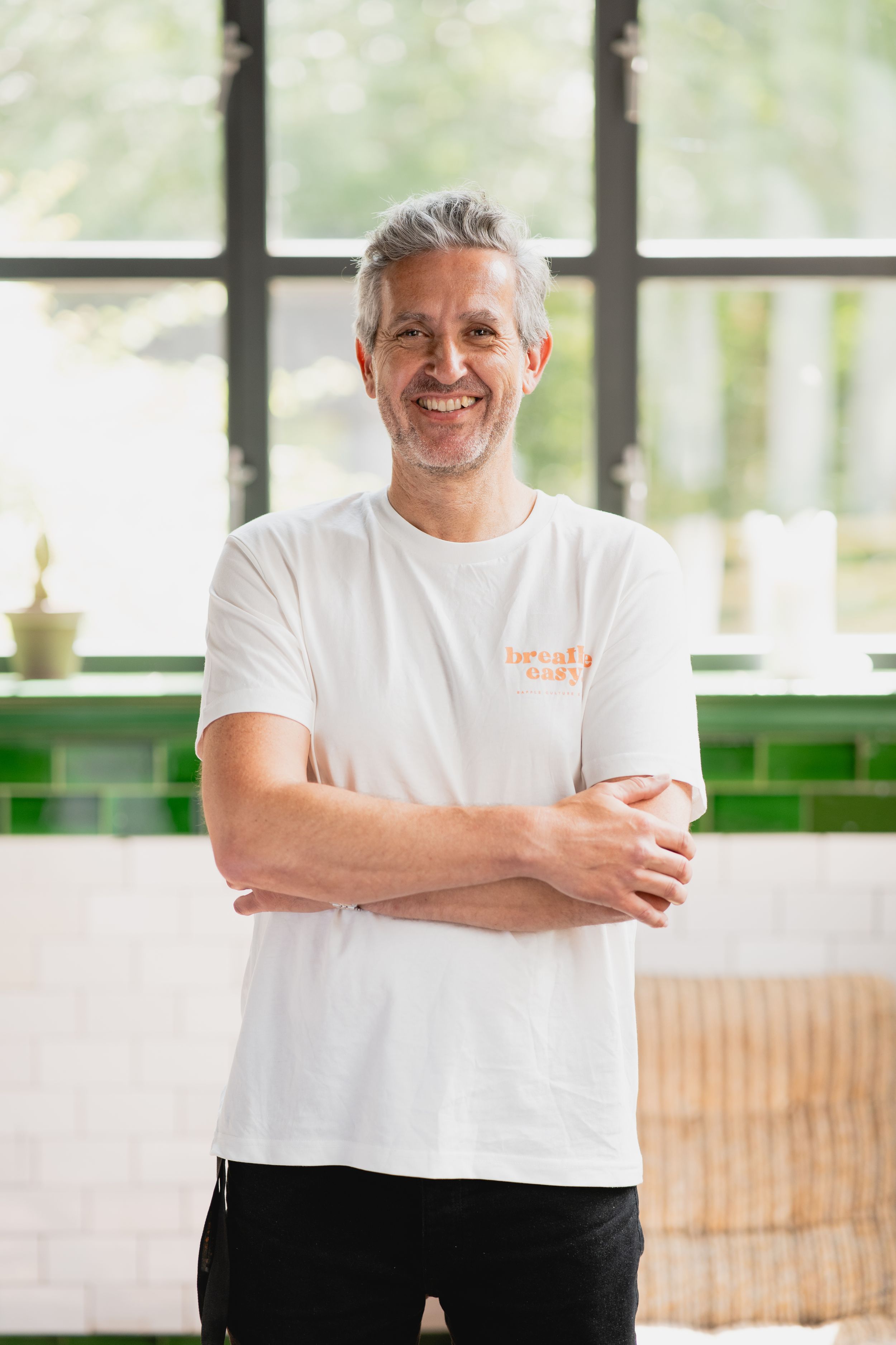 Person in white t-shirt standing in bright office space with crossed arms and warm expression.