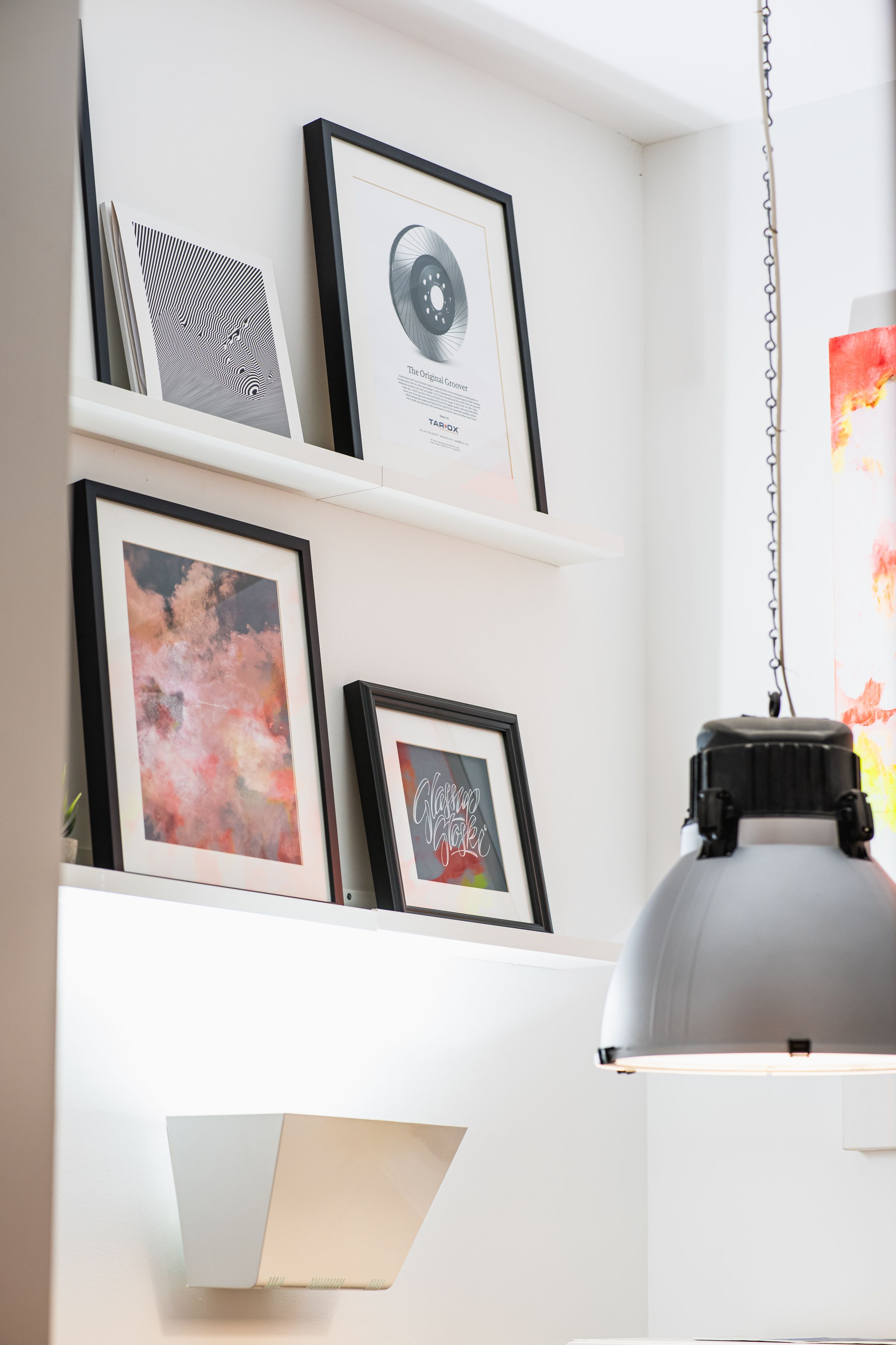 Gallery wall with framed artwork and pendant lights displaying contemporary pieces on white shelves.