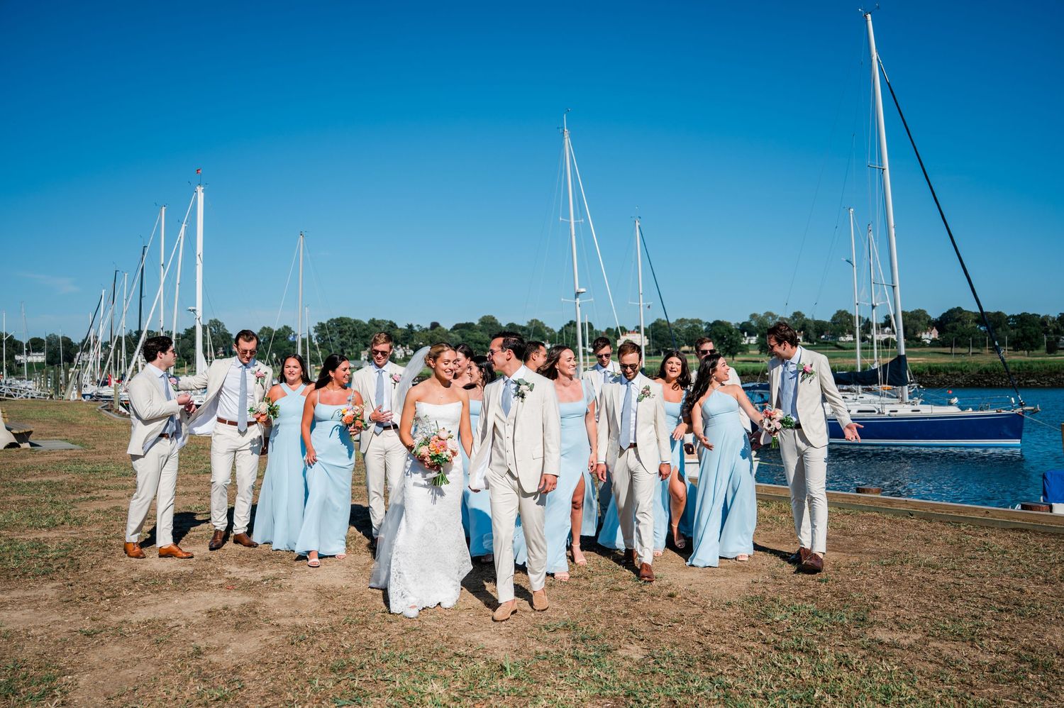 A group in light blue and white attire poses together on a grassy shore with boats and water in the background.